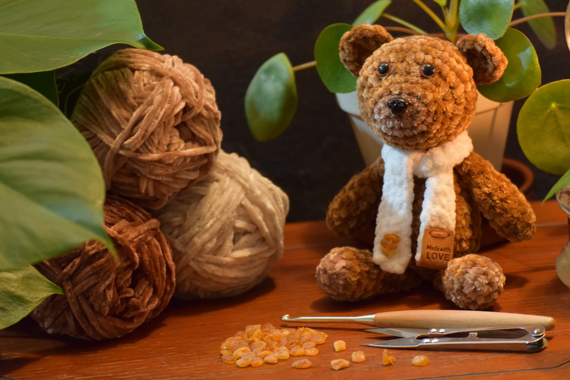 Handmade brown teddy bear with a cream scarf, sitting beside balls of chenille yarn and crochet tools